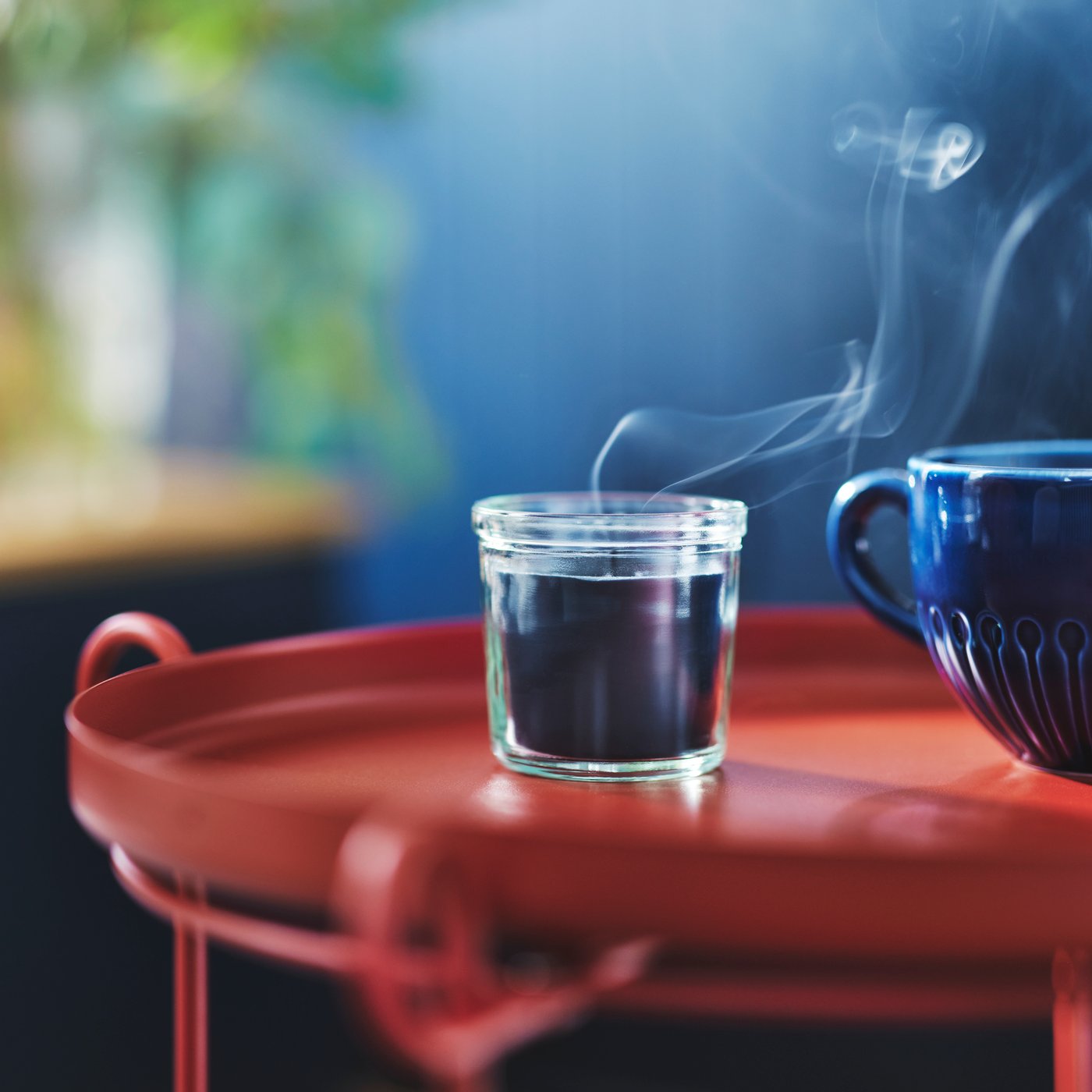 Een rode bijzettafel met daarop een blauwe FRUKTSKOG geurkaars die net is uitgeblazen. Je ziet de rook nog hangen in de lucht.