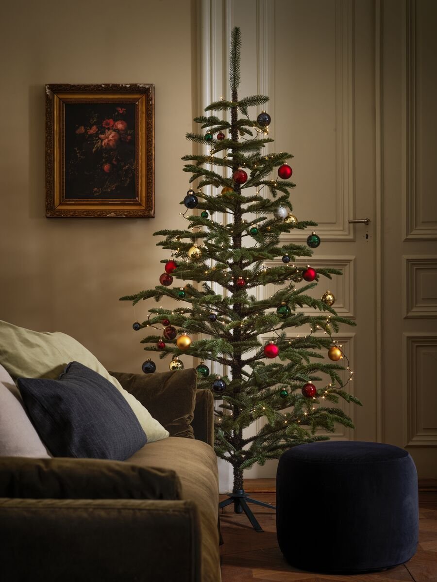 A large holiday tree stands in a traditional-looking living room, its branches decorated with bright-coloured baubles.