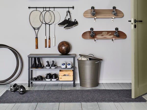 Una banqueta con zapatero PINNIG con zapatillas y zapatos y de fútbol, junto a un basurero KNODD con la tapa abierta. Raquetas y skates están colgados en en la pared de arriba.