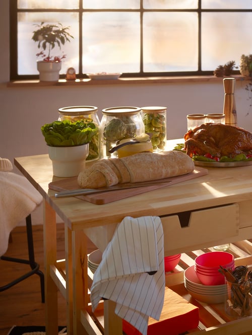 Two solid birch FÖRHÖJA kitchen trolleys placed back-to-back as a wide serving area with jars of food and a loaf of bread.