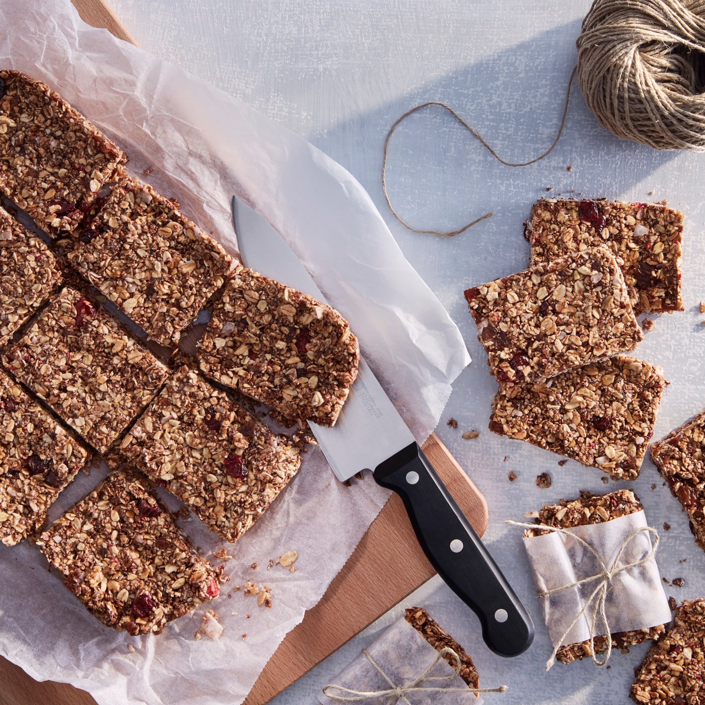 Barras de granola cortadas en forma rectangular sobre un papel de hornear blanco y una tabla de cortar de madera, junto a un cuchillo de cocina y dos trozos envueltos.