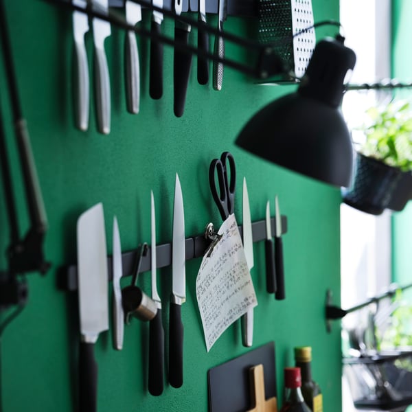 A green wall with different kinds of knives, a pair of scissors, and more attached to a black HULTARP magnet knife rack.