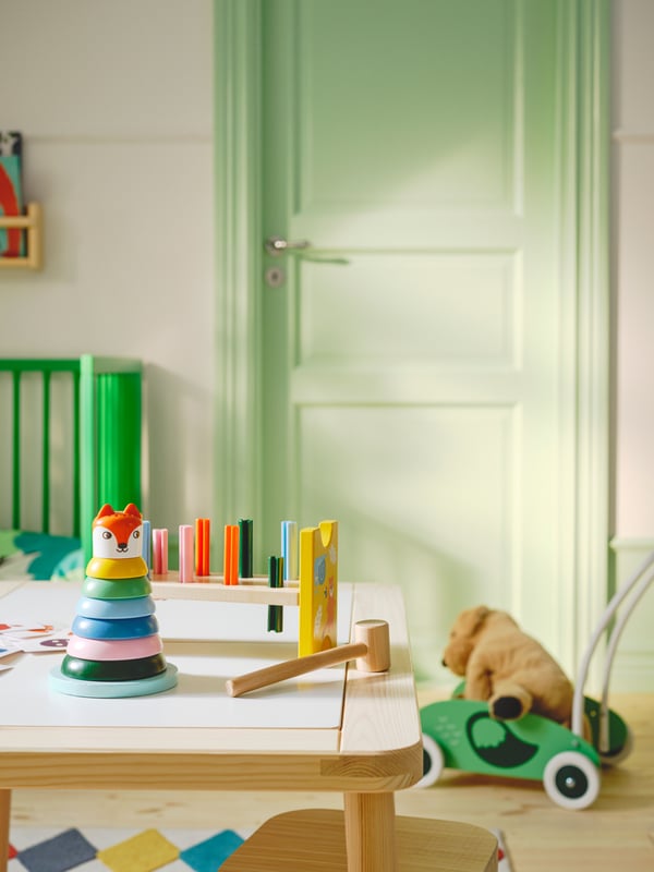 A FLISAT children’s table in a green room with toys on top and a green UPPSTÅ toddler walker on the floor beside it.
