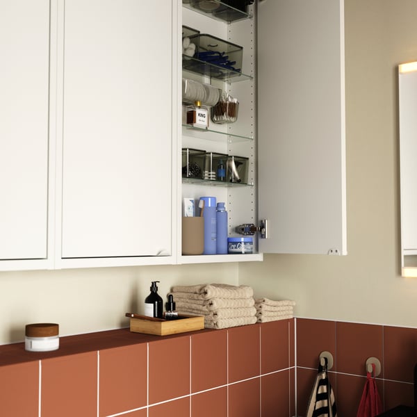 A terracotta-tiled bathroom featuring a white HAVBÄCK wall cabinet, one door open to show glass shelves storing toiletries.