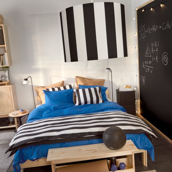 A black and white striped RINGSTA lamp shade hanging with a bed in the background made with blue, brown and striped bedlinen.