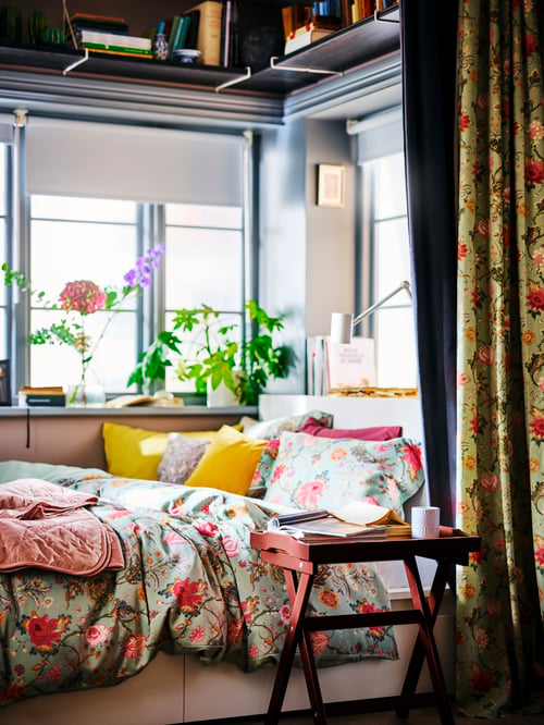 Living room with a white bed frame with two storage boxes, a tray table, a green fabric, a white wall lamp, cushion covers.