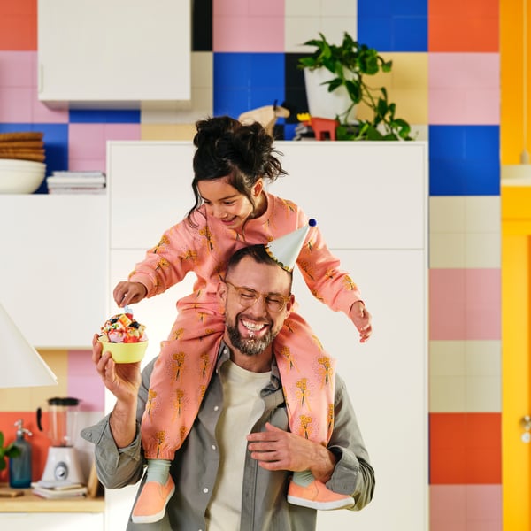 A grown-up with a party hat is holding a KALAS bowl with sweets. A child sitting on their shoulders reaches for the sweets.
