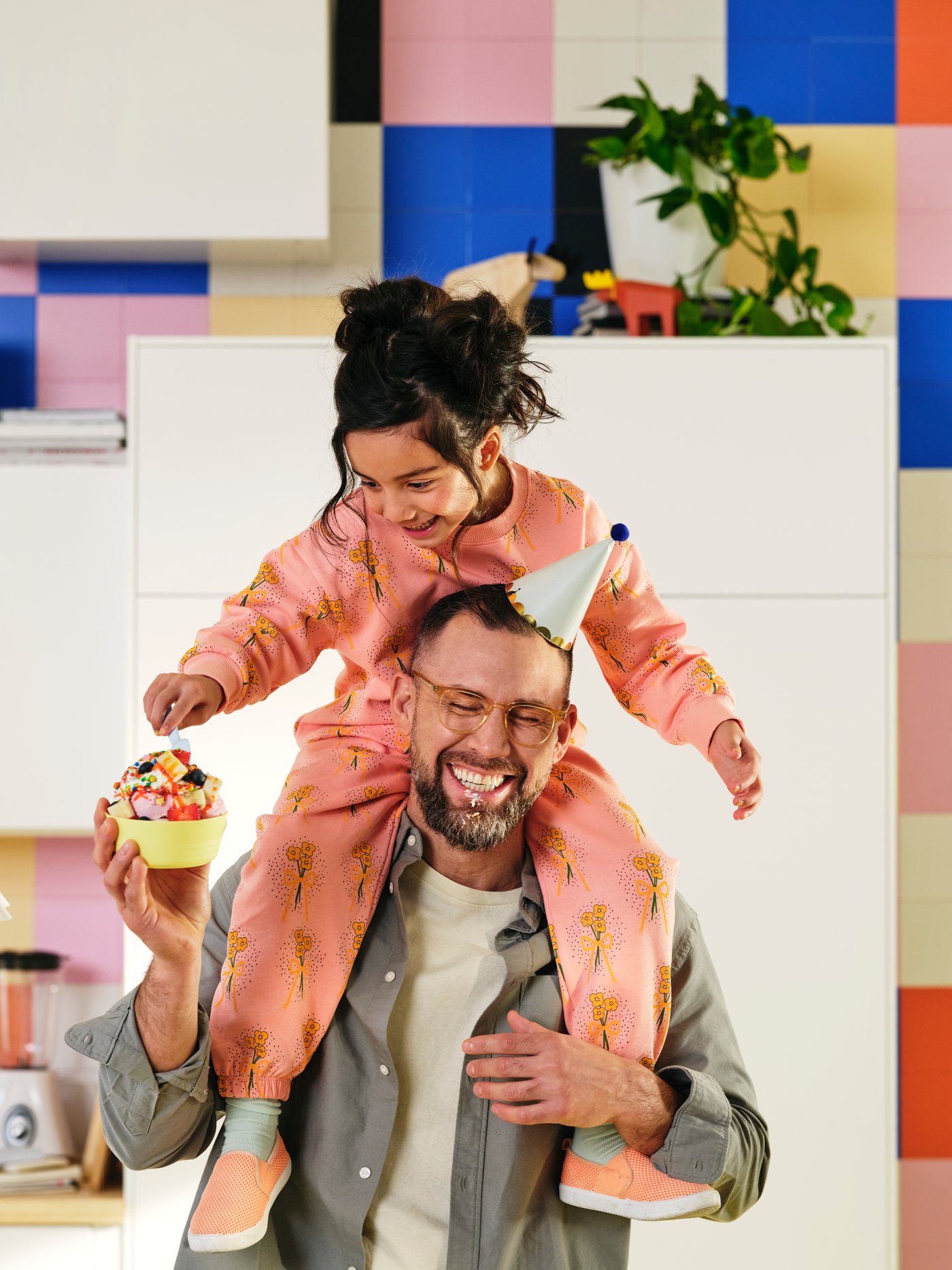 A grown-up with a party hat is holding a KALAS bowl with sweets. A child sitting on their shoulders reaches for the sweets.