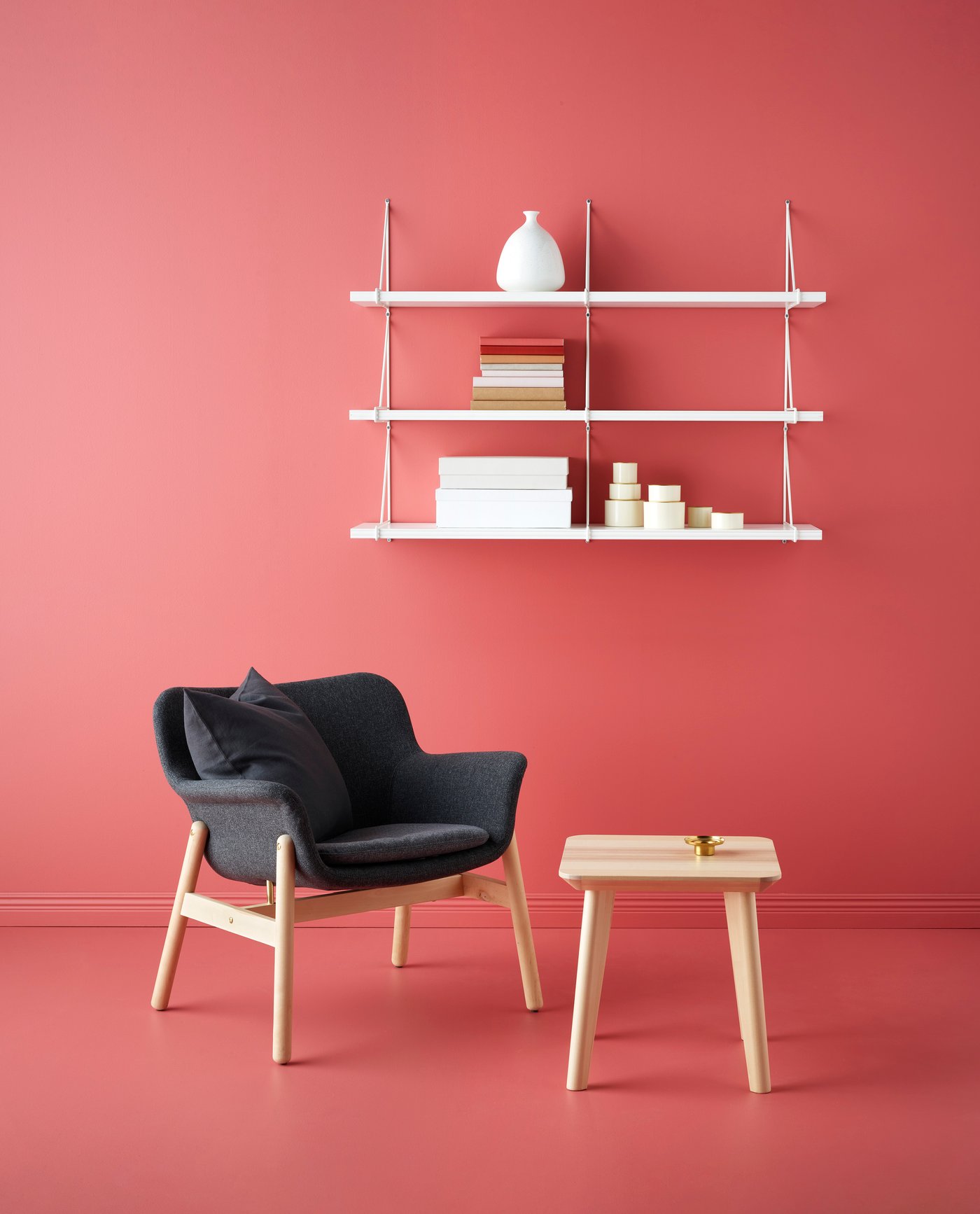 Modern inredning med röd vägg och golv, mörkgrå fåtölj med träben, litet sidobord och tre vita vägghyllor med böcker, ljus och dekorationer.