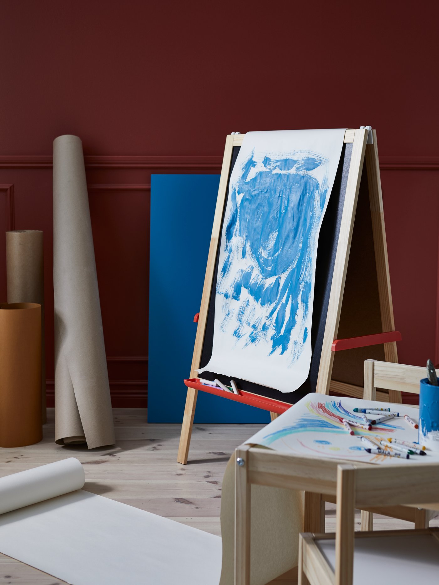 Una habitación con un caballete MÅLA en mitad de un proceso de pintura y un rollo de papel de dibujo de la misma colección usado como lienzo, rodeado de accesorios de manualidades.