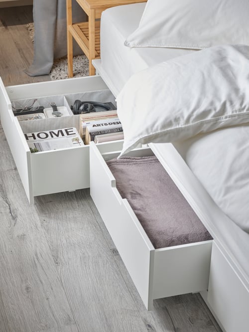 Modern bedroom featuring a bed frame with six drawers, two of each are open revealing a bedspread and magazines.