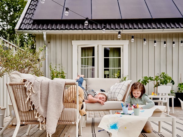 Hai dous nenos nun patio. Un está tombado nun sofá, mentres que o outro colorea nunha mesa. O patio está unido a unha casa cun teito de paneis solares.