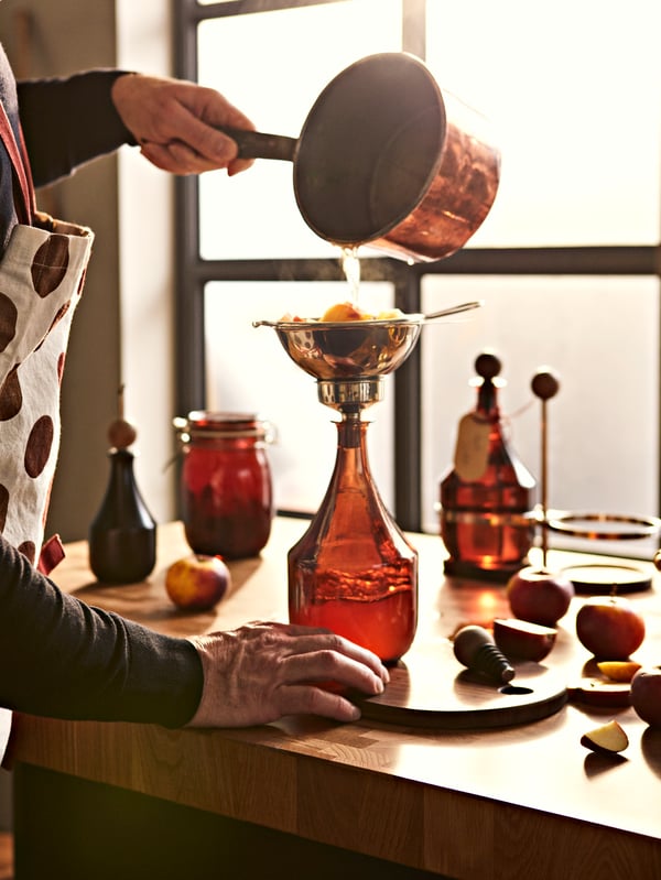 A man is in the kitchen using the KRÖSAMOS strainer and funnel set over a KRÖSAMOS glass bottle to make an apple vinaigrette.