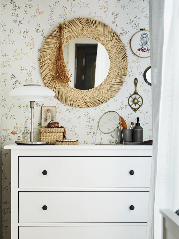 A VRIGSTAD mirror on a floral-papered wall above a white HEMNES chest with 3 drawers, on which stands a TÄLLBYN table lamp.