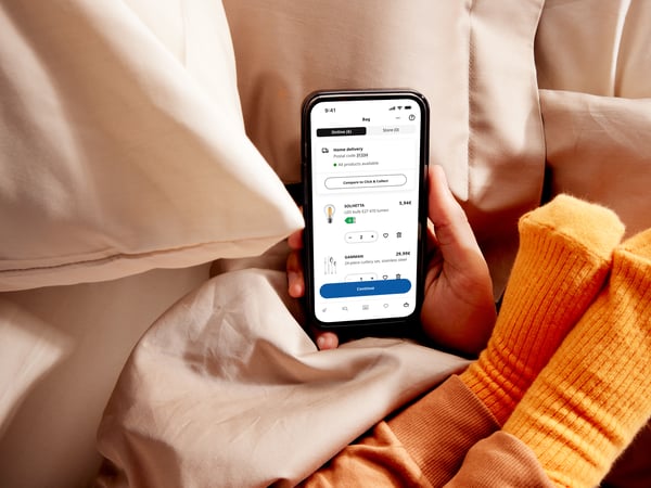 On a bed, a hand is holding a smartphone displaying the IKEA app, a small child's feet with orange socks rests on the persons arm.
