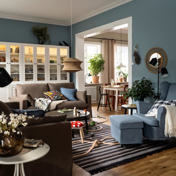 Two HYLTARP sofas stand opposite a STRANDMON wing chair and footstool on a woollen RÅVAROR rug in a flexible living room.