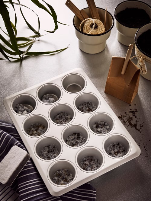 Blacha do pieczenia muffinów VARDAGEN. W większości jej otworów nasiona na wilgotnym papierze. Na blacie papierowy worek i nasiona.