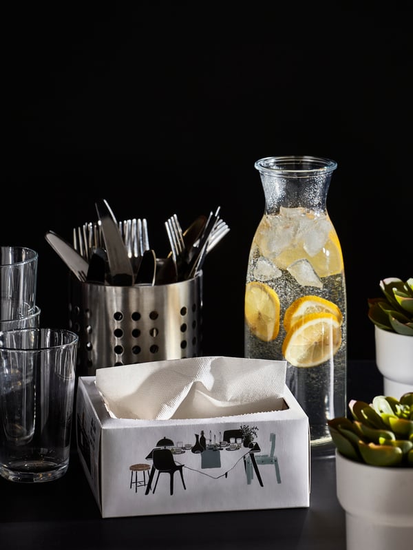 Drinking glasses, cutlery, a carafe with water and slices of lemon, a box of white paper napkins and two small plants.