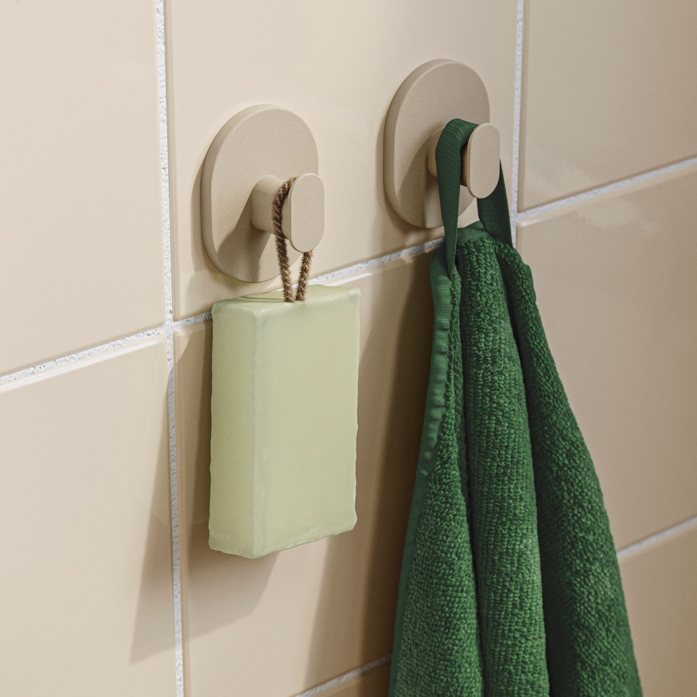A close-up of a set of 2 grey-beige FLORSJÖN hooks on a tiled bathroom wall, with a green towel and a mint green bar of soap.