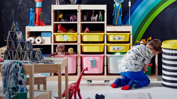 Un niño jugando en el suelo con un peluche. En la habitación se ven muebles con juguetes, una mesa LILLABO en madera con un castillo de naipes encima.
