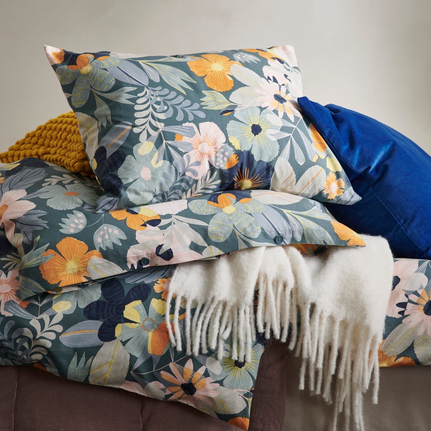 A pile of FÄLTGRÄSMOTT, SANELA and SVARTPOPPEL cushions and a KLIPPOXEL white throw placed on a brown floor.