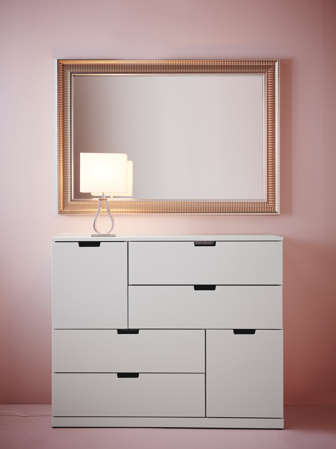 Dormitorio con paredes y suelo de color rosa, cómoda blanca de seis cajones, lámpara de mesa niquelada, espejo plateado en la pared.