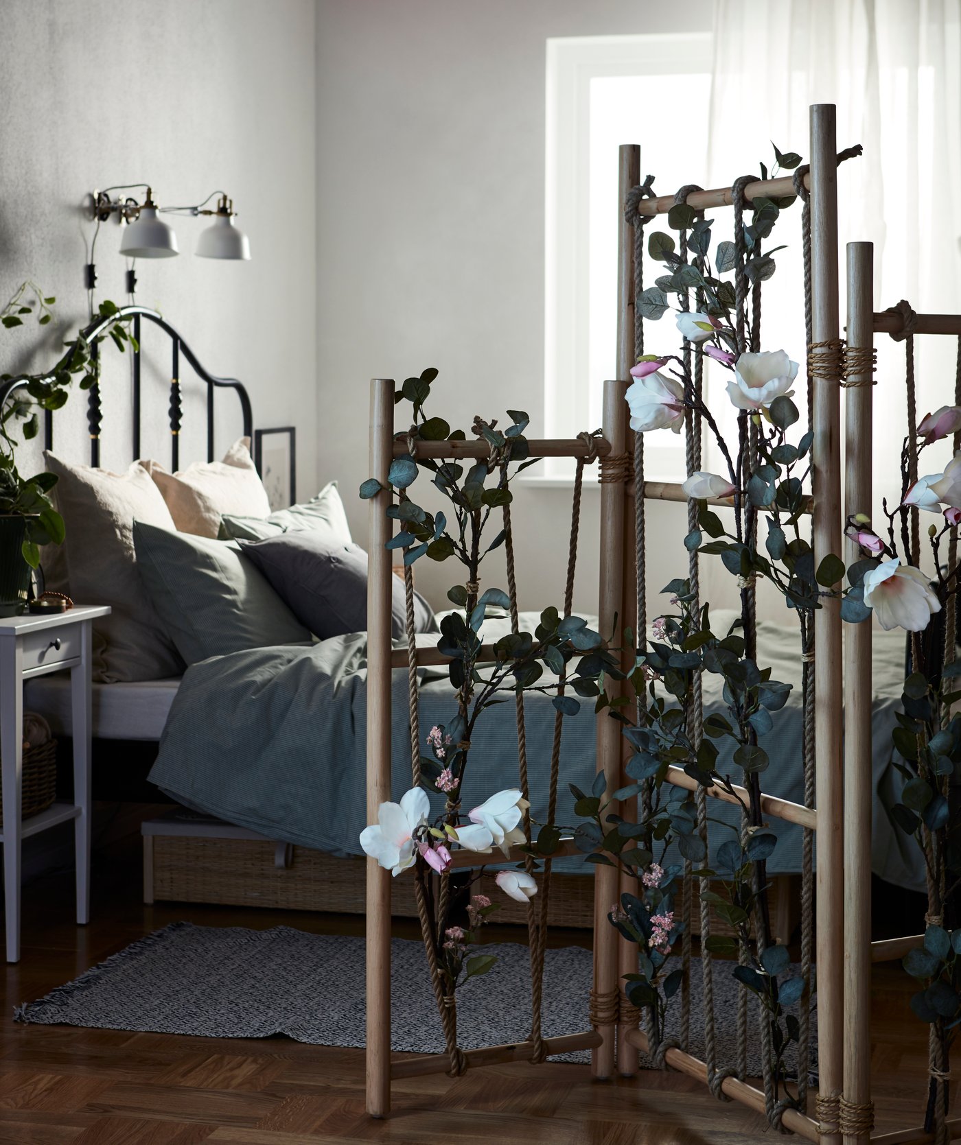 Schlafzimmer, in dem das Bett durch einen TÄNKVÄRD Raumteiler vom Rest des Raums abgetrennt wird. TÄNKVÄRD ist mit Kunstblumen geschmückt.