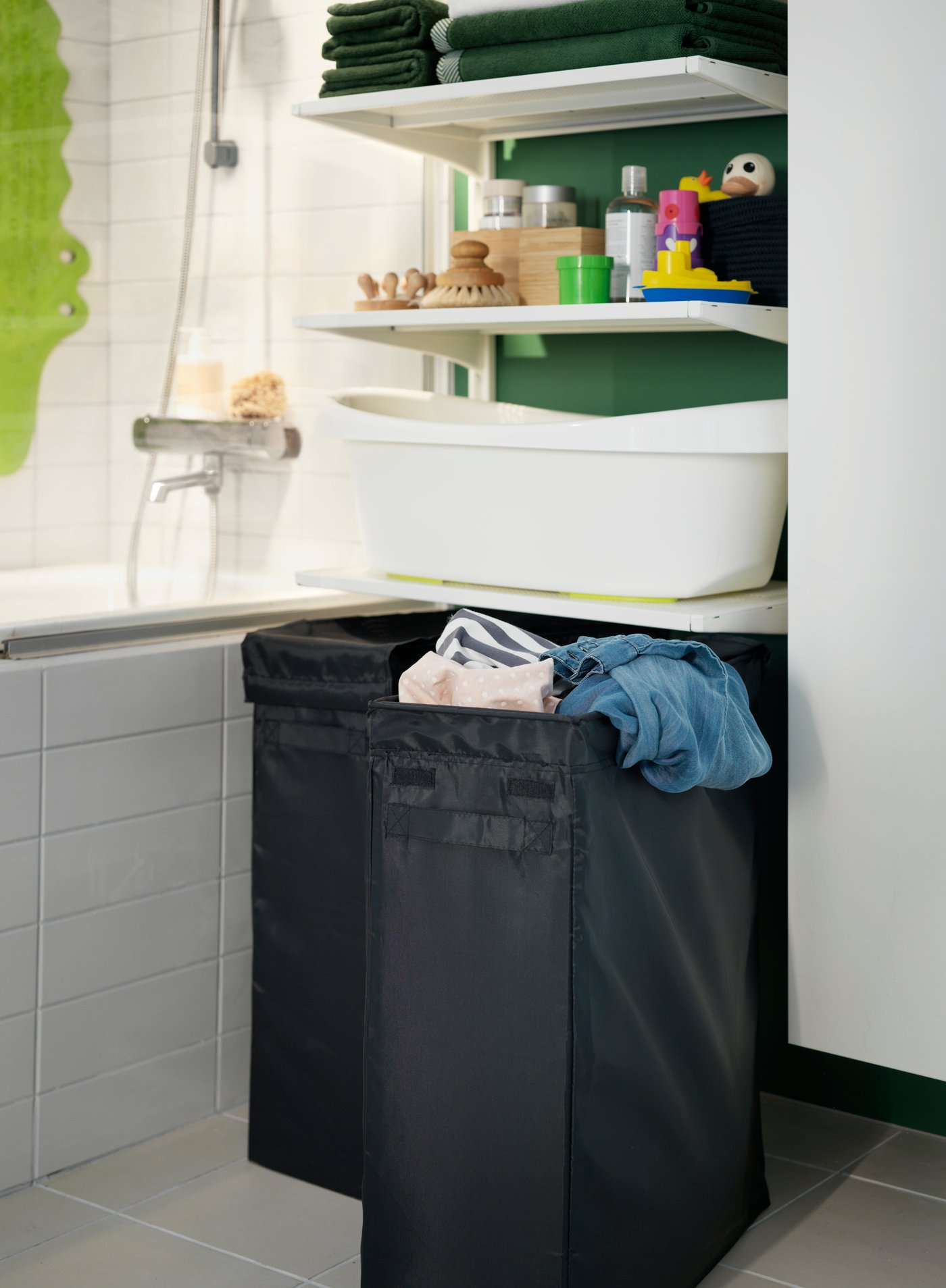 Detalle del espacio inferior entre un mueble y una tina de baño donde, bajo un conjunto de repisas blancas con accesorios de baño, hay dos canastos de ropa sucia SKUBB negros. 