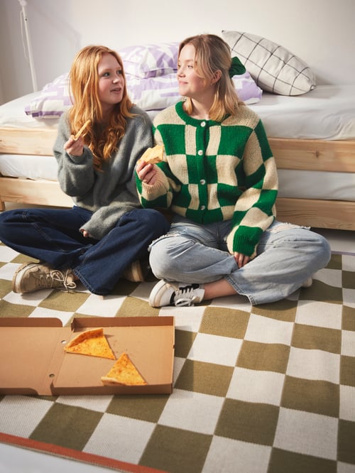Două persoane care dețin felii de pizza stau pe un covor cu carouri KLASSRUM și pe marginea unui pat UTÅKER.