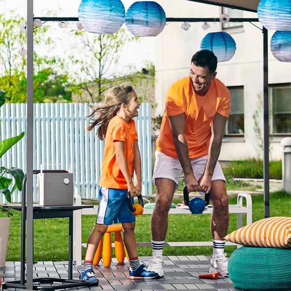 Un uomo e un bambino che fanno una allenamento con alcuni accessori da palestra all'aperto in un giardino. A lato, un pouf verde con un cuscino a righe.