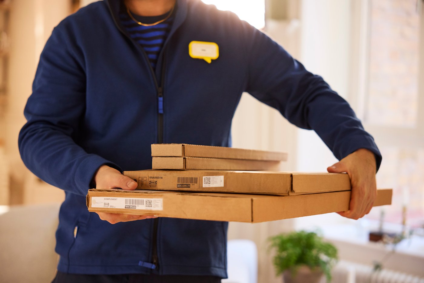 Person in blue work clothes holds several flat packages of furniture parts in his hands in an indoor environment.
