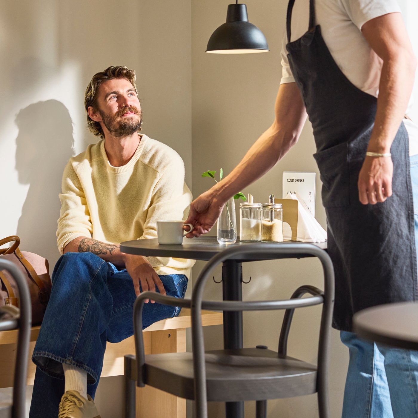 Barista serverar kaffe vid ett litet kafébord medan den sittande kunden tittar upp; koppar, sockerburkar och varmt ljus skapar en mysig miljö.