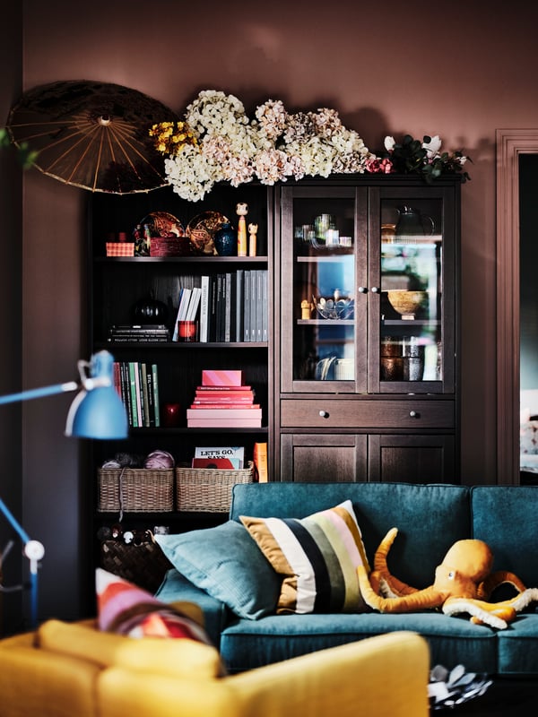 A black-brown HEMNES glass-door cabinet showing wine glasses and dinnerware, standing in front of a green Sofa.