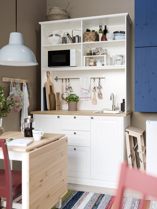 A ÖNNERUP kitchenette with a microwave oven a kettle, and a mug is placed next to a rack with knobs and a gateleg table.