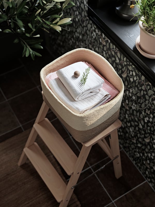 Folded white and pink towels are placed in a LJUNGAN basket sitting on a step stool next to a window and some green plants.