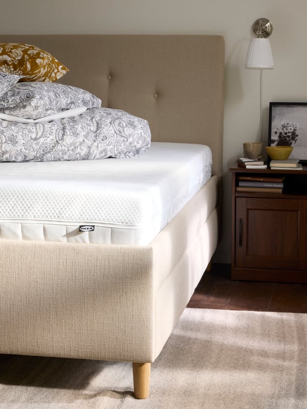 IDANÄS upholstered bed frame in beige with white mattress visible and the bedding folded in a pile on top of the bed.