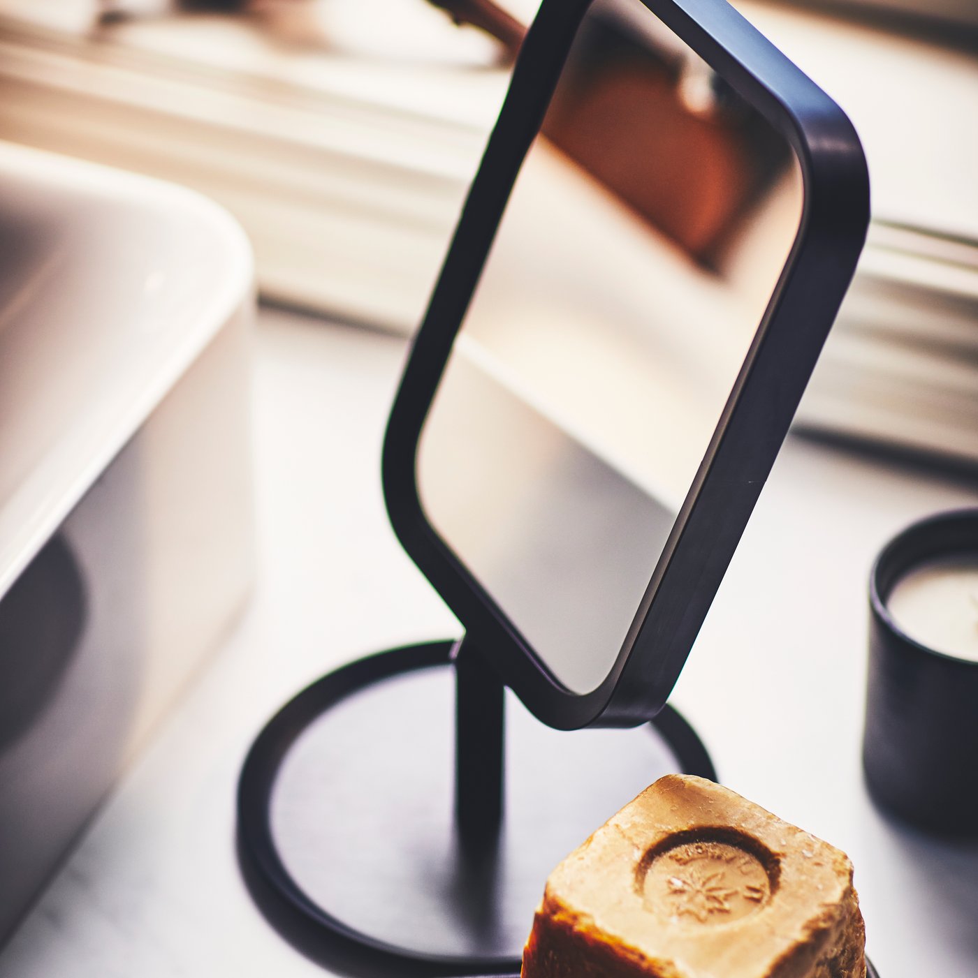 Un miroir de table LINDBYN noir et un pain de savon posés sur un meuble pour lavabo GODMORGON gris foncé, près d’un lavabo, sous un rebord de fenêtre.