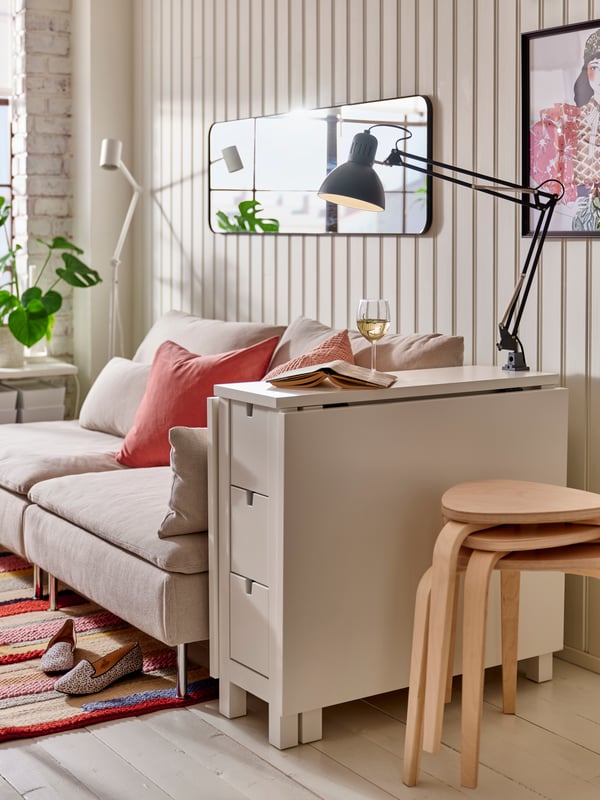 A white NORDEN gateleg table with a dark grey work lamp, beside a light beige sofa with cushions and a stack of birch stools.