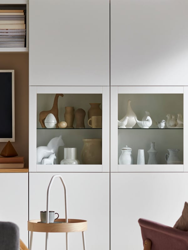 A white BESTÅ with two glass doors filled with different kinds of ornaments. In front of the cabinet is a birch BURVIK side table.