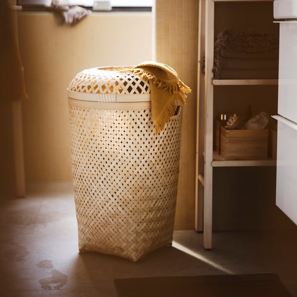 A bathroom with a bamboo SALUDING laundry basket with lid, folded bath towels, a wooden storage unit, toiletries and boxes.
