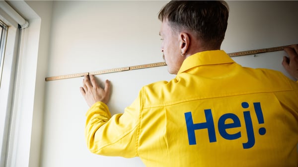 An IKEA co‑worker wearing a yellow jacket with “Hej!” printed on the back measures and marks a wall inside a home interior.
