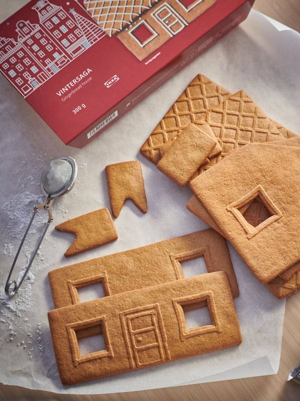 Parts of a VINTERSAGA gingerbread house are spread out on a piece of baking paper, next to the red VINTERSAGA packaging.