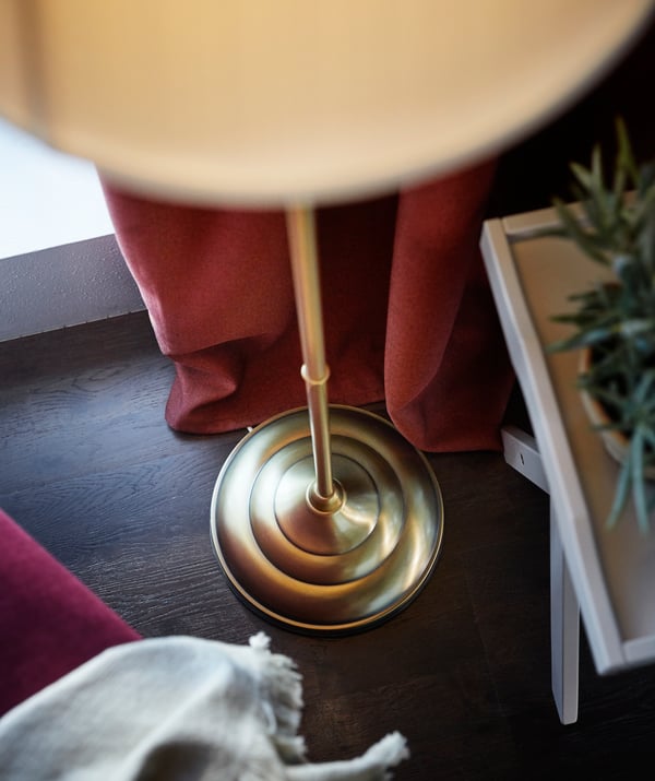 Detailansicht eines goldenen Leuchtenfuß einer ÅRSTID Stehlampe. Im Hintergrund ein roter Vorhang und ein weißes Tischtablett.