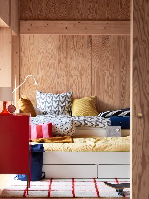 Ein Teenagerzimmer mit einem weißen SLÄKT Bettgestell, Kissen und Bettwäsche in verschiedenen Farben, einem rotweißen Teppich und einer Tasche.