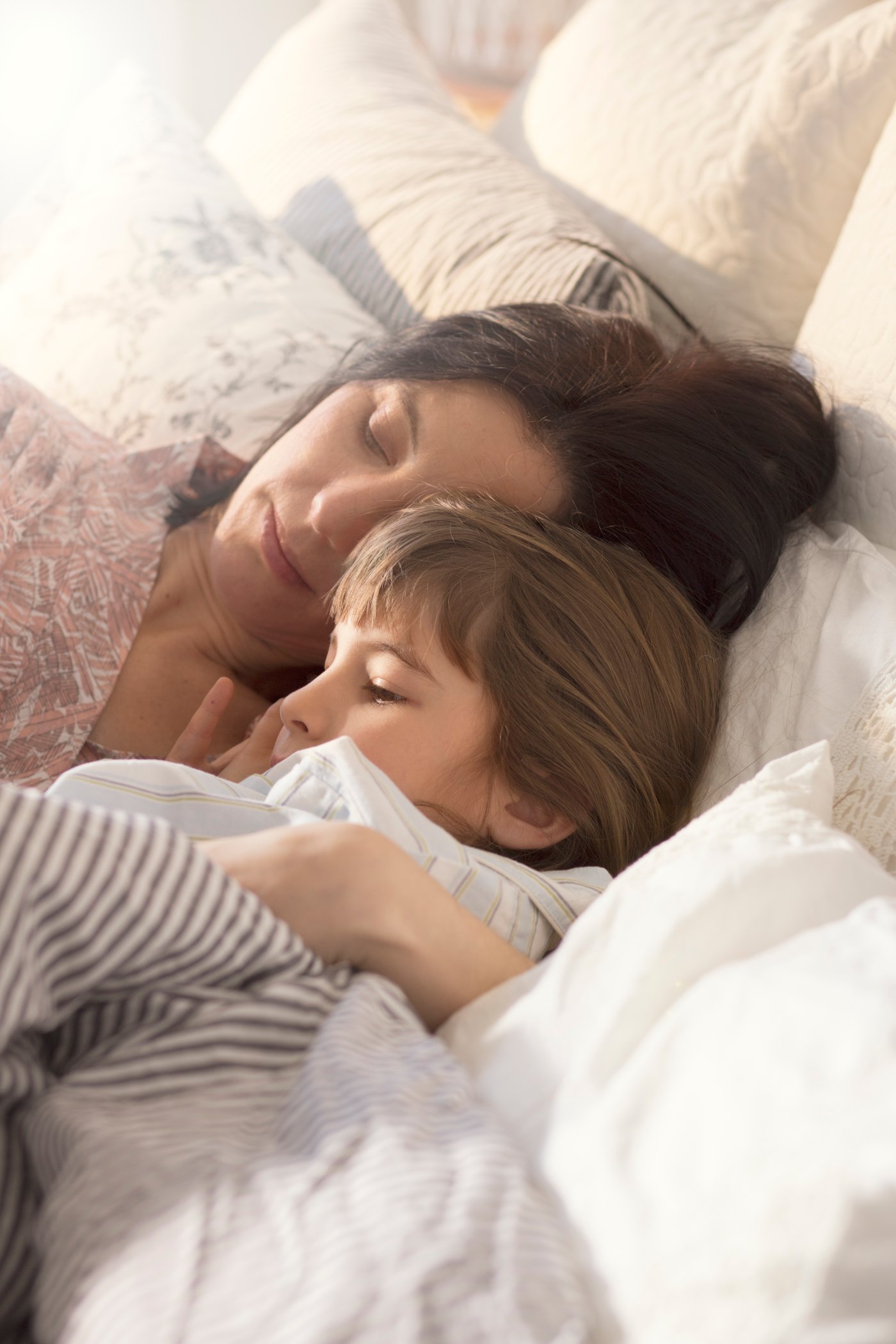 En kvinde ligger i en seng og holder om et barn de er begge i pyjamas og ligger på puder og med dynen over sig.