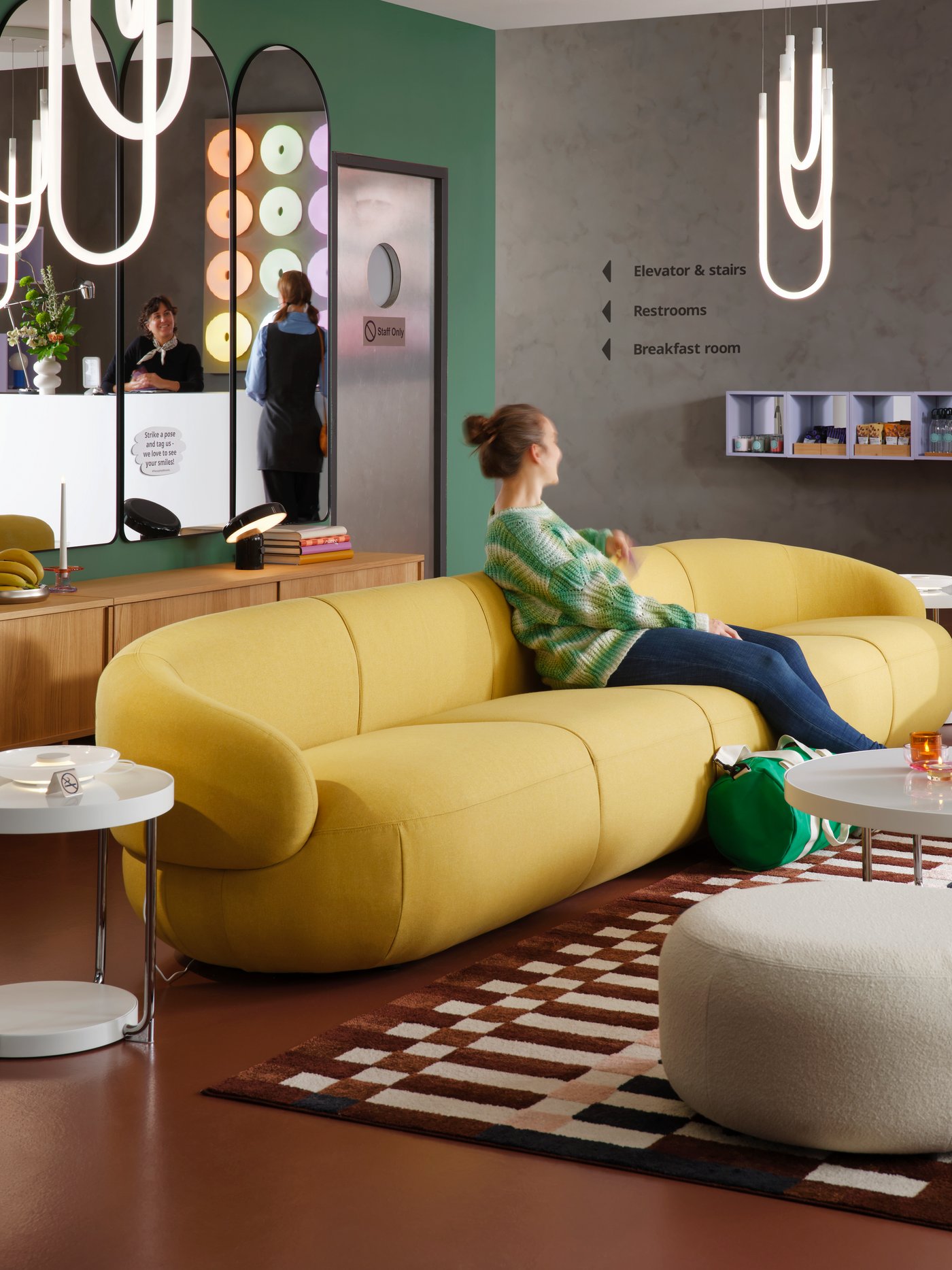 A multicolour, check patterned MARKBLÅMME low-pile rug is in a modern hotel lounge that has guests on the sofa and at reception.