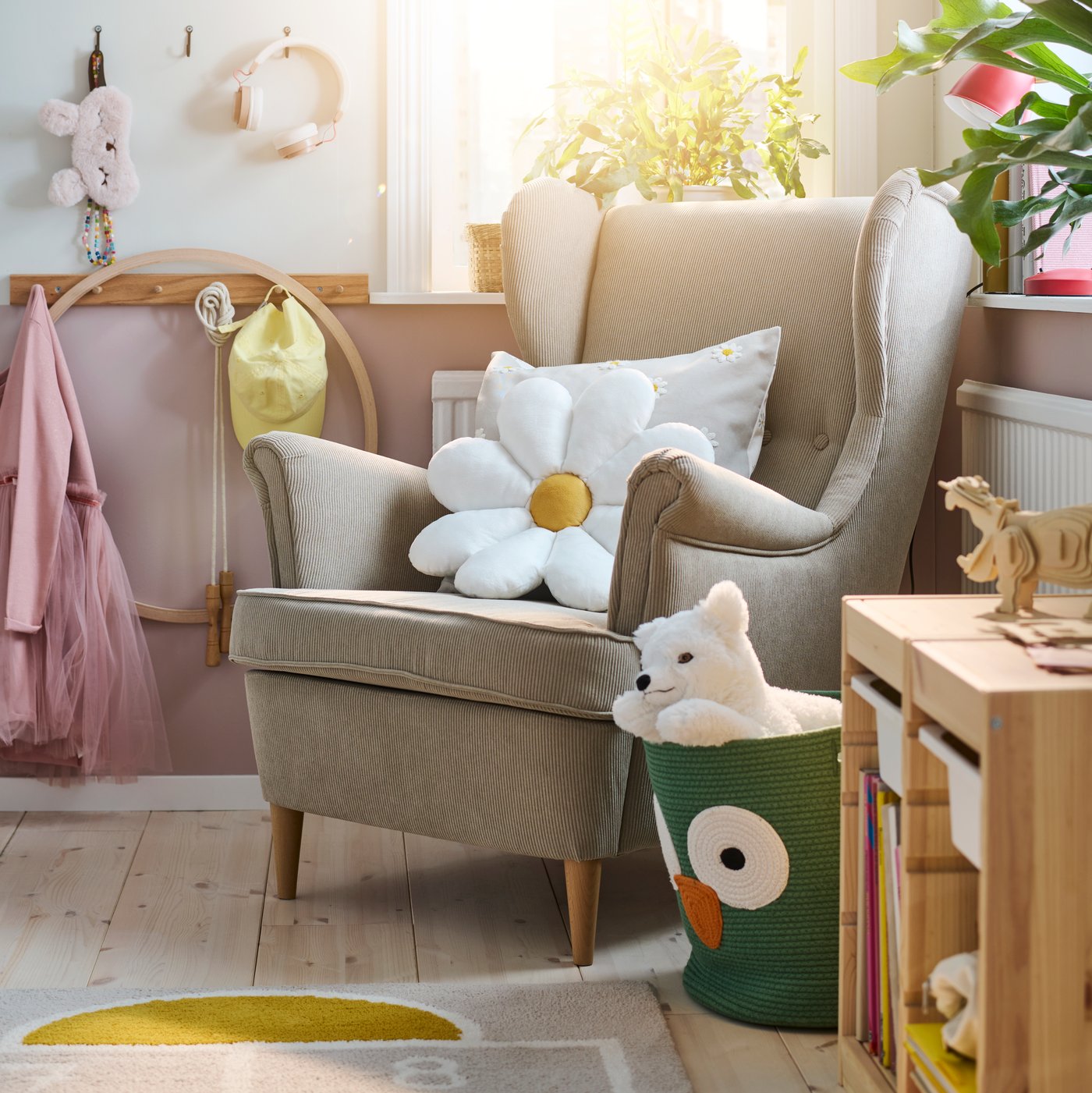 A beige STRANDMON wing chair in a bright children’s bedroom with a white flower-shaped SKOGSDUVA cushion on it.
