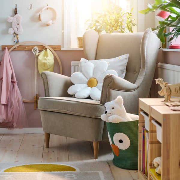 Unha cadeira de brazos con orelleiras STRANDMON beixe nun dormitorio infantil luminoso cun coxín branco SKOGSDUVA con forma de flor enriba.