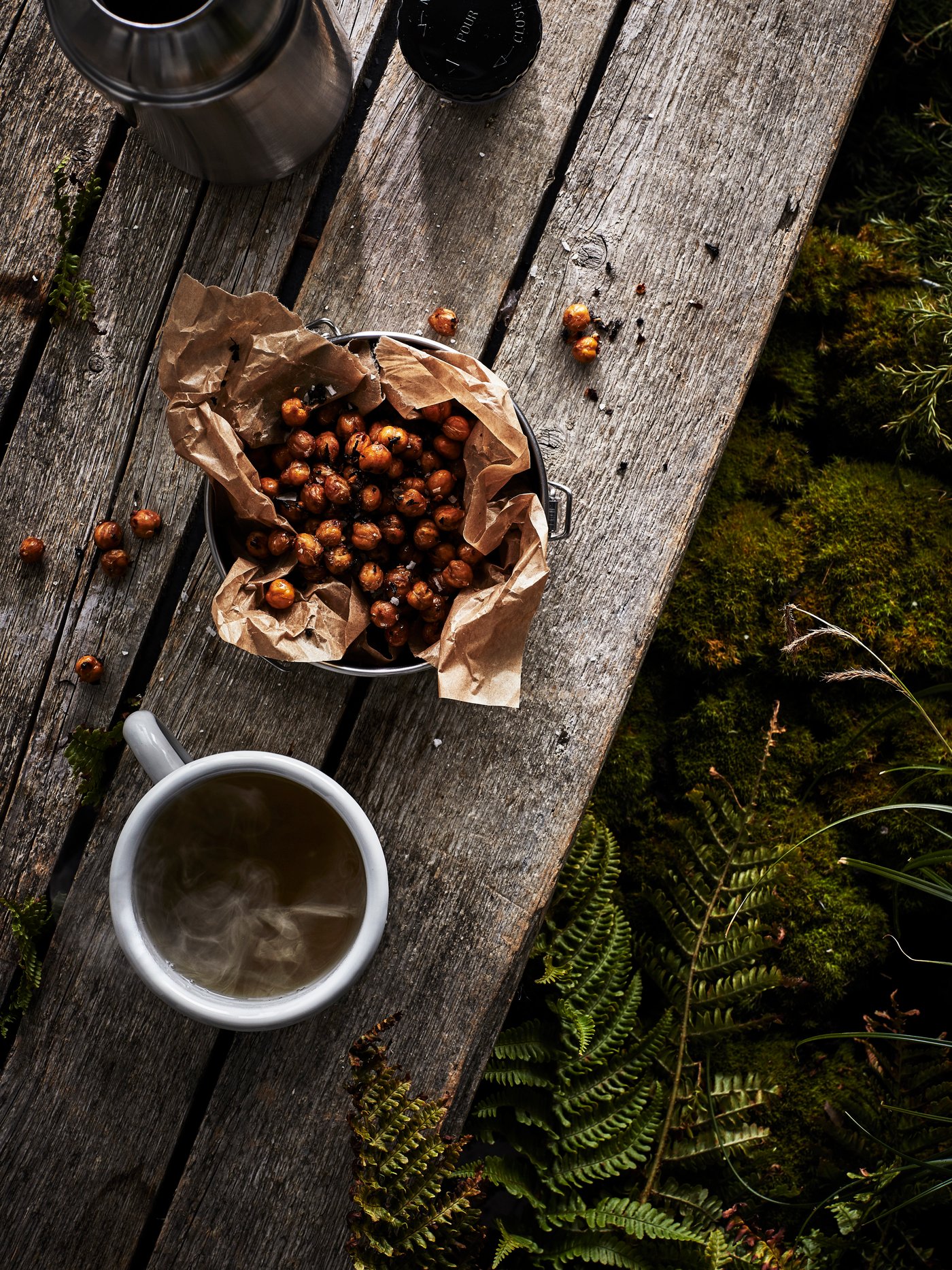 A cup of EGENTID tea beside a bowl of chai and chilli roasted chickpeas and an open thermos flask on a wooden table.
