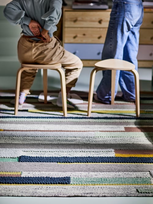 A multicolour BRÖNDEN rug with two birch stools on top, with one person standing and another sitting and storage behind.